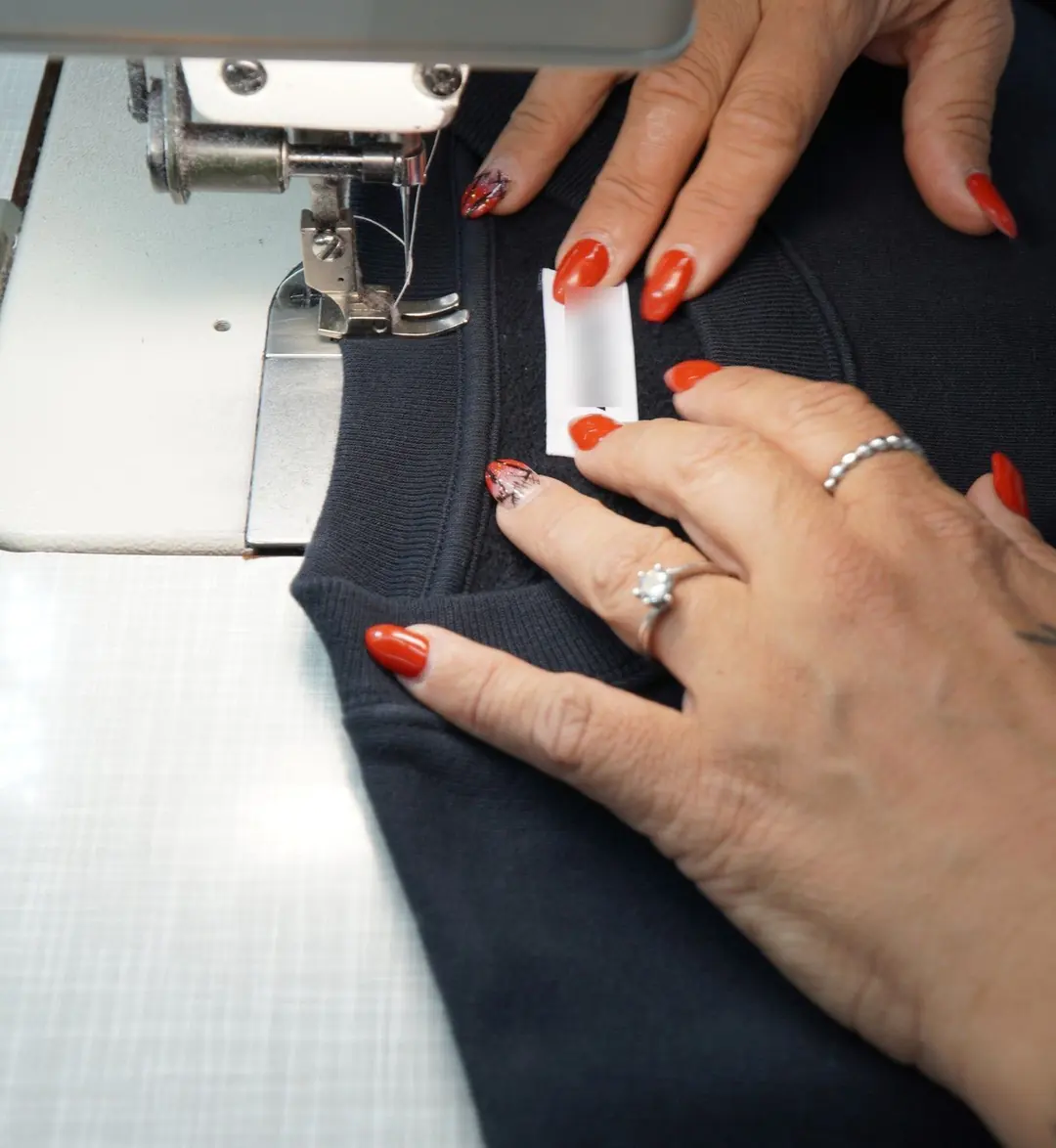 Hands guiding a sweatshirt under a sewing machine as a neck label is stitched in
