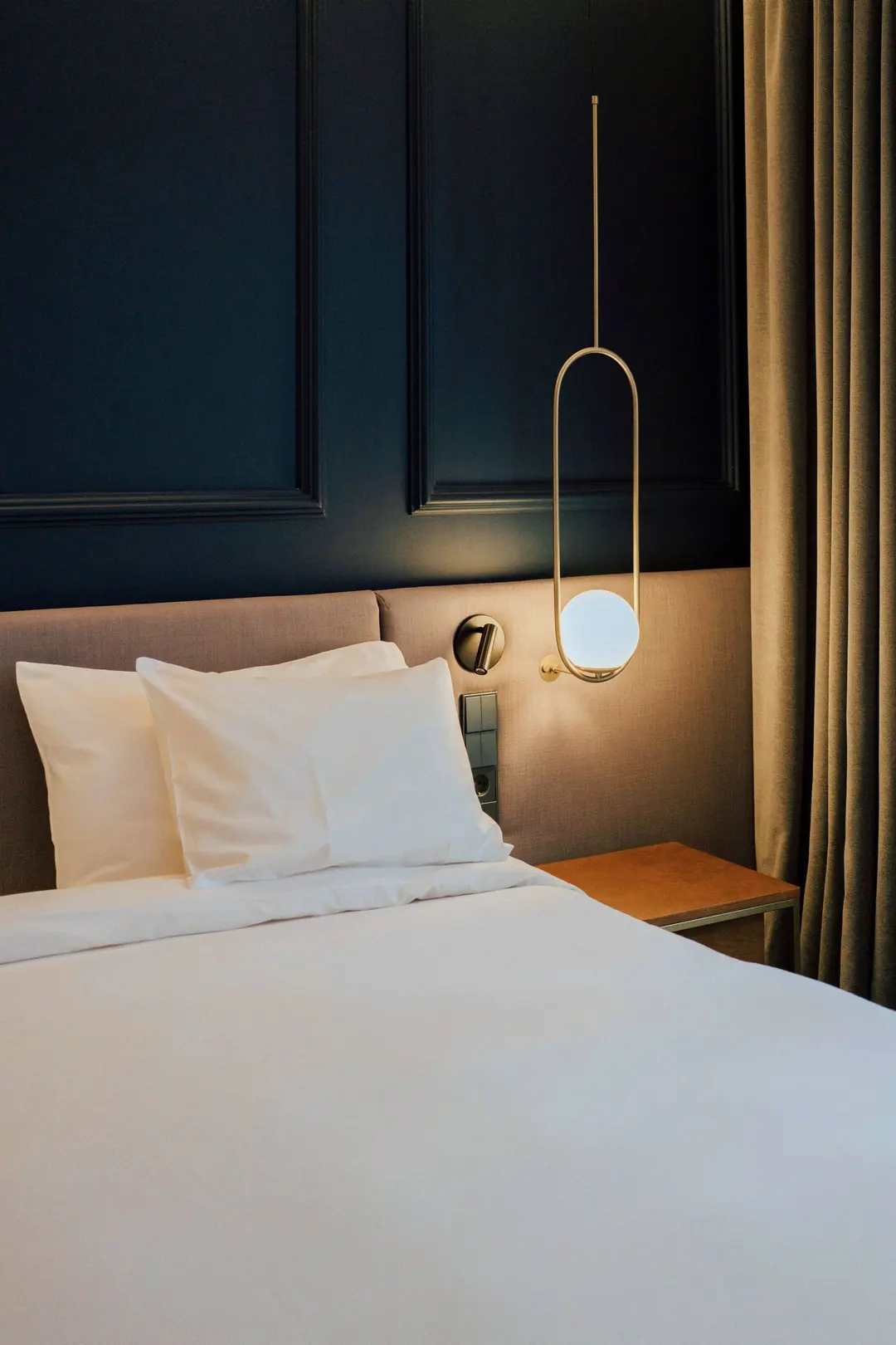Hotel bed with white linen against a dark blue wall and a brass pendant lamp