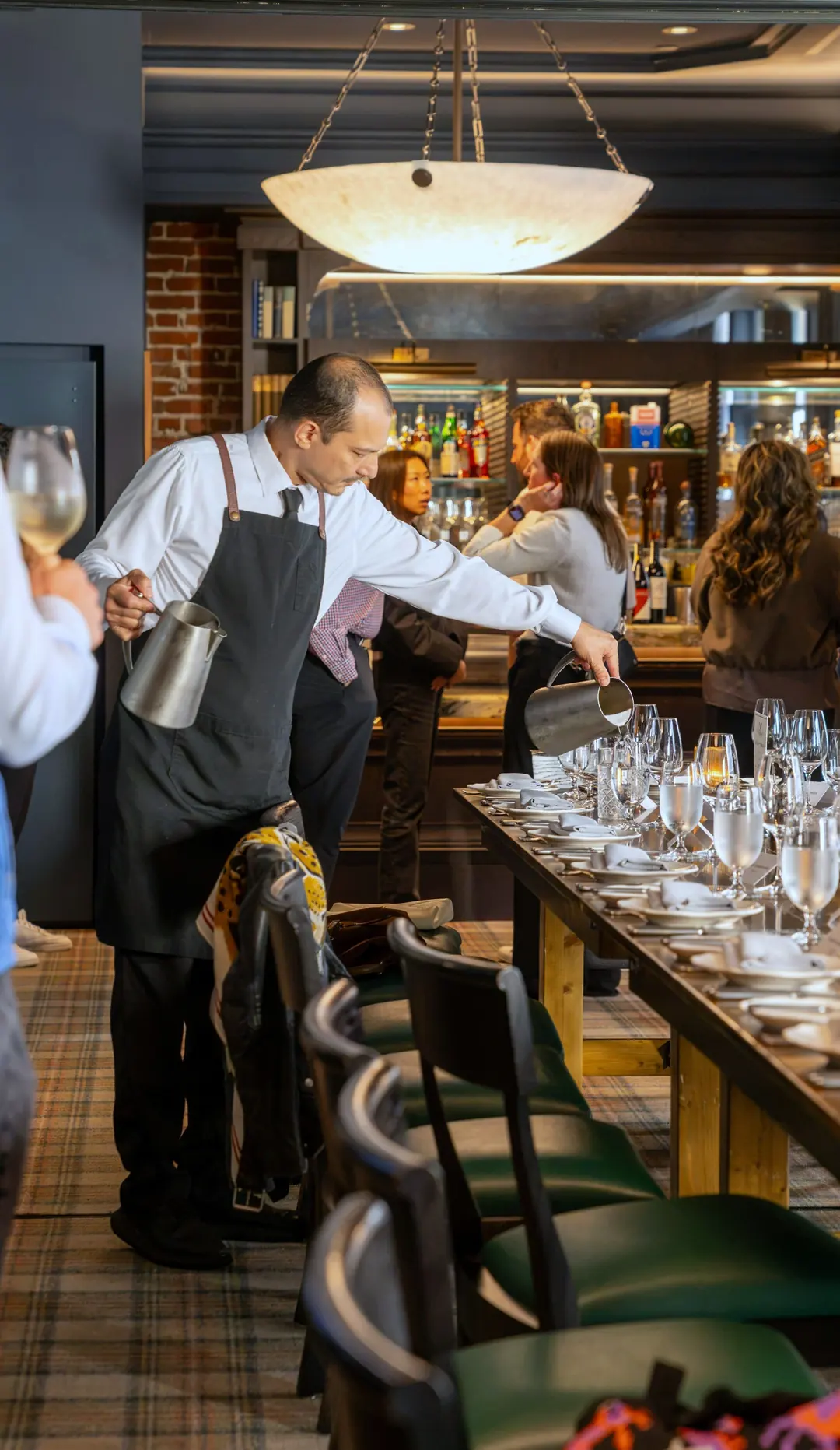 Server in a white shirt, tie and long dark apron pouring water along a laid restaurant table